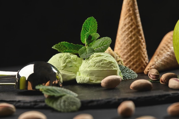 Inquadratura ravvicinata di un gelato al pistacchio piccante decorato con menta, coni di cialda con pistacchi sparsi si trovano nelle vicinanze, servito con una paletta di metallo su un'ardesia di pietra su uno sfondo nero.