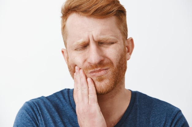 Inquadratura ravvicinata di un uomo dai capelli rossi con la barba che sente dolore ai denti, aggrottando le sopracciglia e facendo espressione di sofferenza con gli occhi chiusi, toccando la guancia, avendo bisogno di chiamare il dentista per curare la carie o il dente marcio