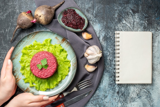 Insalata di barbabietola vista dall'alto sul piatto nelle mani della donna barbabietole all'aglio barbabietola grattugiata in una piccola ciotola forchetta e coltello scialle viola blocco note sul tavolo grigio