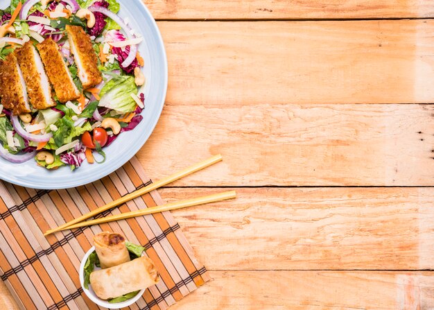 Insalata di filetto fritto con involtini primavera in ciotola di ceramica e bacchette contro tavolo in legno