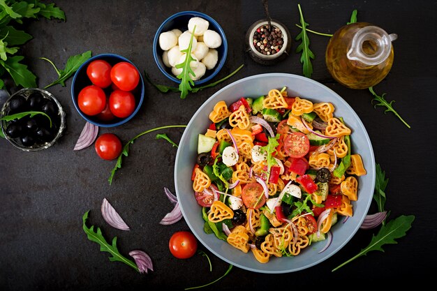 Insalata di pasta a forma di cuore con pomodori, cetrioli, olive, mozzarella e cipolla rossa alla greca. Disteso. Vista dall'alto