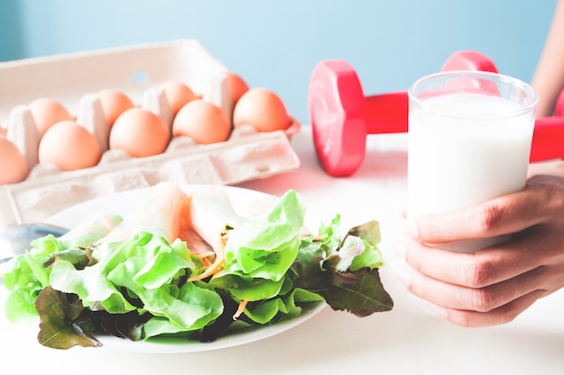 Insalata fresca con uovo e mano che tiene vetro di latte, menu sano con dumbbells rossi, concetto di stile di vita sano