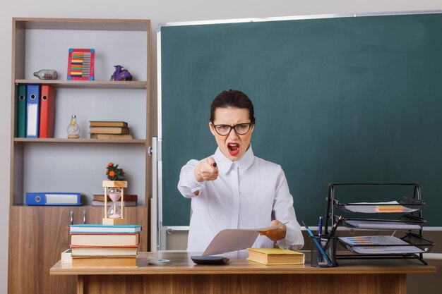 Insegnante di giovane donna che indossa occhiali controllando i compiti degli studenti arrabbiati e frustrati che puntano alla telecamera seduta al banco di scuola davanti alla lavagna in classe