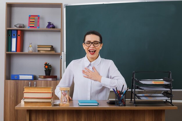 Insegnante di giovane donna che indossa occhiali sorridendo allegramente tenendo la mano sul petto sentendosi grato seduto al banco della scuola davanti alla lavagna in classe