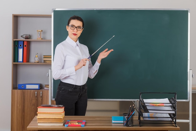 Insegnante di giovane donna che indossa occhiali tenendo il puntatore mentre spiega la lezione guardando fiducioso in piedi al banco della scuola davanti alla lavagna in classe