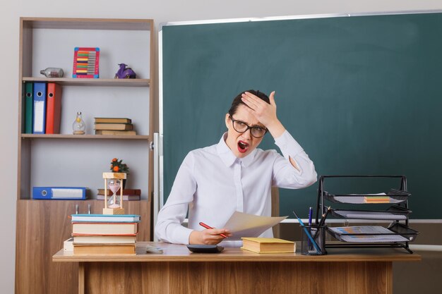 Insegnante di giovane donna con gli occhiali che controlla i compiti degli studenti che sembrano confusi con la mano sulla fronte ha dimenticato qualcosa seduto al banco di scuola davanti alla lavagna in classe