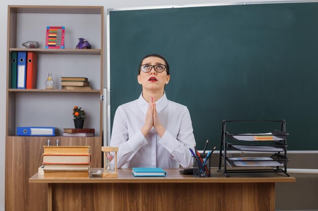 Insegnante di giovane donna con gli occhiali che sembra triste tenendo i palmi delle mani insieme come pregare seduto al banco di scuola davanti alla lavagna in classe