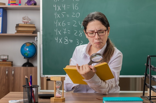 Insegnante di giovane donna con gli occhiali che tiene libro guardando le pagine attraverso la lente di ingrandimento con espressione fiduciosa seduto al banco di scuola davanti alla lavagna in aula