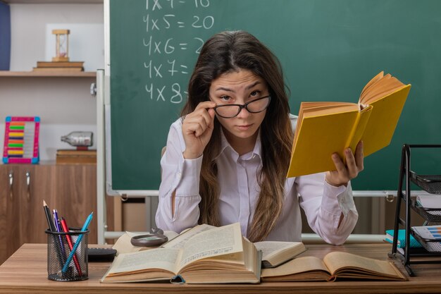Insegnante di giovane donna con gli occhiali con il libro è concentrato seduto al banco di scuola davanti alla lavagna in aula