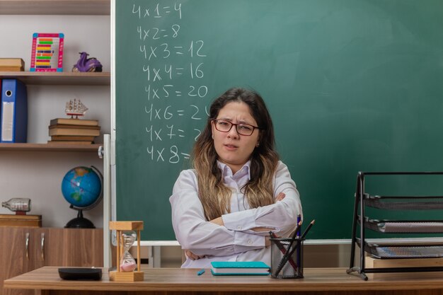 Insegnante di giovane donna con gli occhiali guardando davanti confuso e scontento con le braccia incrociate seduto al banco di scuola davanti alla lavagna in aula