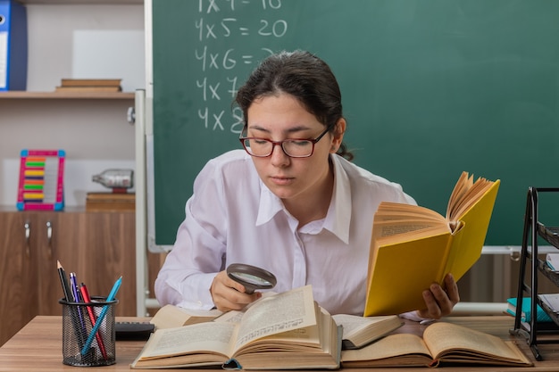 Insegnante di giovane donna con gli occhiali guardando il libro attraverso la lente di ingrandimento essendo confuso e dispiaciuto seduto al banco di scuola davanti alla lavagna in aula