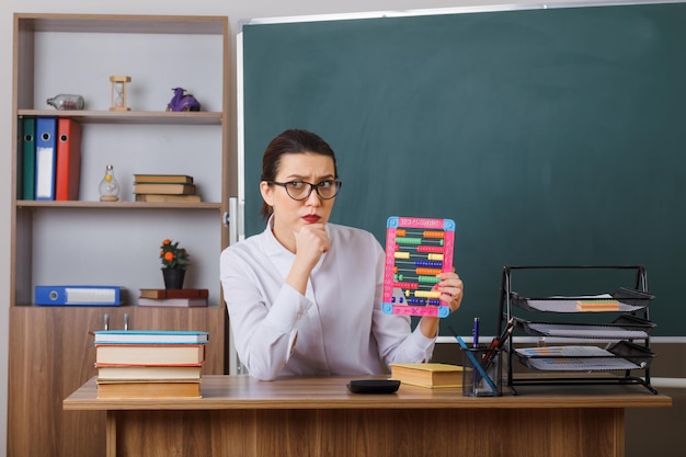 Insegnante di giovane donna con gli occhiali in possesso di abaco che spiega la lezione guardando con espressione pensierosa pensando seduto al banco della scuola davanti alla lavagna in classe