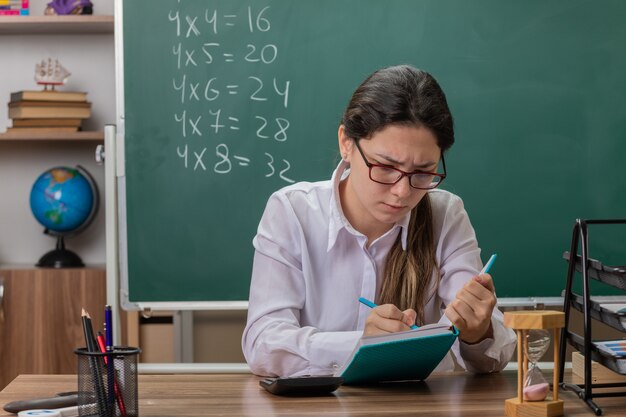 Insegnante di giovane donna con gli occhiali scrivendo qualcosa in notebook preparando per la lezione guardando fiducioso seduto al banco di scuola davanti alla lavagna in aula