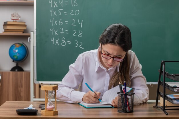 Insegnante di giovane donna con gli occhiali scrivendo qualcosa in notebook preparando per la lezione guardando fiducioso seduto al banco di scuola davanti alla lavagna in aula