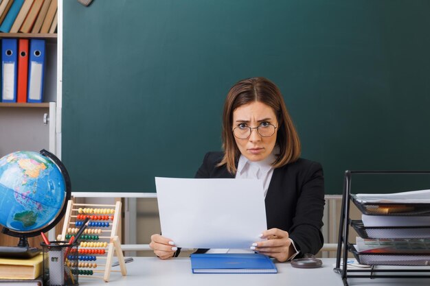 Insegnante di giovane donna con gli occhiali seduto al banco della scuola con il globo e i libri davanti alla lavagna in classe con in mano un foglio di carta bianco vuoto guardando la fotocamera accigliata