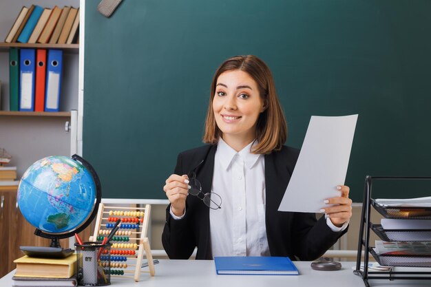 Insegnante di giovane donna con gli occhiali seduto al banco della scuola con il globo e i libri davanti alla lavagna in classe con un foglio di carta bianco vuoto felice e sorridente positivo