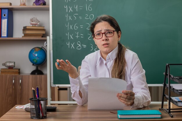 Insegnante di giovane donna con gli occhiali seduto al banco di scuola con pagine bianche che sembrano confusi e scontenti davanti alla lavagna in aula