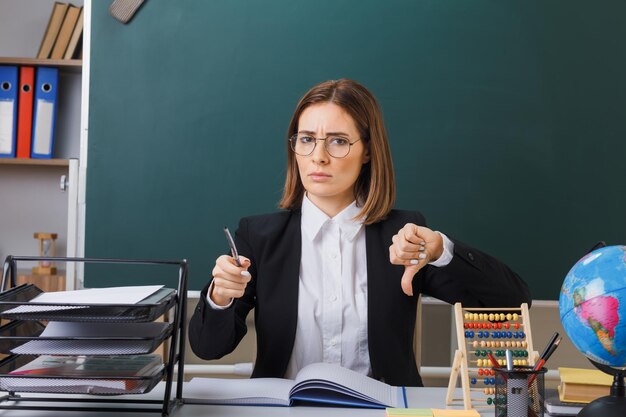 Insegnante di giovane donna con gli occhiali seduto al banco di scuola davanti alla lavagna in classe con abaco e globo controllando il registro di classe tenendo il puntatore che mostra il pollice verso il basso accigliata