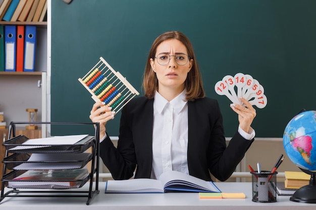 Insegnante di giovane donna con gli occhiali seduto al banco di scuola davanti alla lavagna in classe con targhe e abaco che spiegano la lezione guardando confuso e deluso