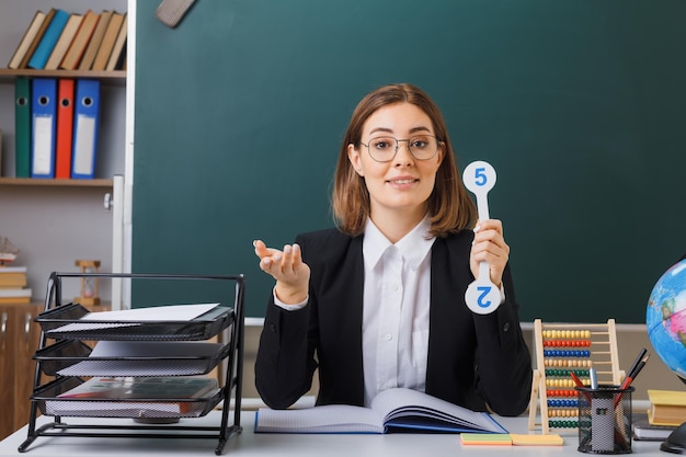 Insegnante di giovane donna con gli occhiali seduto al banco di scuola davanti alla lavagna in classe tenendo le targhe spiegando la lezione alzando il braccio come per fare la domanda