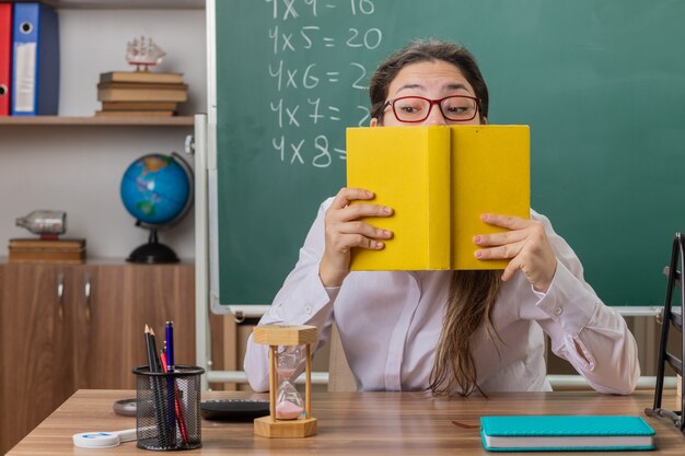 Insegnante di giovane donna con gli occhiali tenendo il libro in preparazione per la lezione di essere confuso e preoccupato seduto al banco di scuola davanti alla lavagna in aula