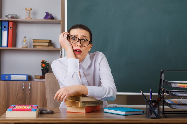 Insegnante di giovane donna con occhiali che controllano i compiti degli studenti che guardano la fotocamera confusi e sorpresi seduti al banco della scuola davanti alla lavagna in classe