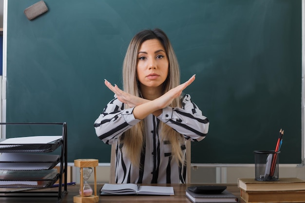 insegnante di giovane donna seduta al banco di scuola davanti alla lavagna in classe controllando i compiti degli studenti che guardano la fotocamera con la faccia seria incrociando le mani maing gesto di arresto