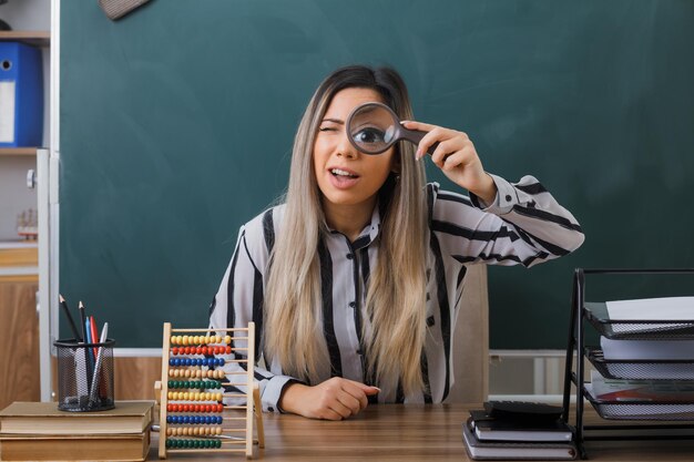 Insegnante di giovane donna seduto al banco di scuola davanti alla lavagna in classe spiegando la lezione guardando la fotocamera attraverso la lente d'ingrandimento che sembra sorpreso