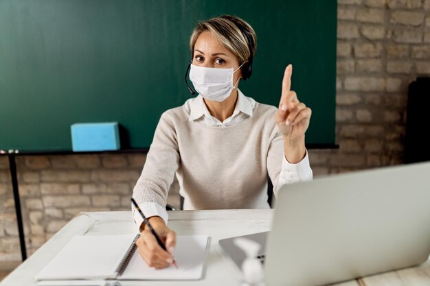 Insegnante di scuola che indossa una maschera facciale e tiene una lezione online dall'aula a causa dell'epidemia di coronavirus.