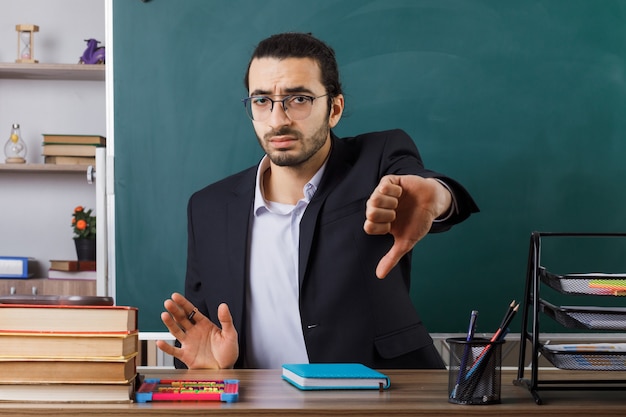 Insegnante di sesso maschile che mostra il pollice verso il basso con gli occhiali seduto al tavolo con gli strumenti della scuola in classe