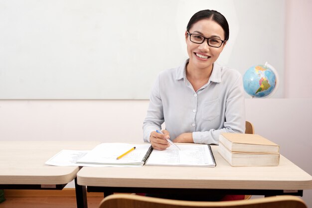 Insegnante femminile asiatica sorridente che si siede allo scrittorio in aula e che scrive in taccuino