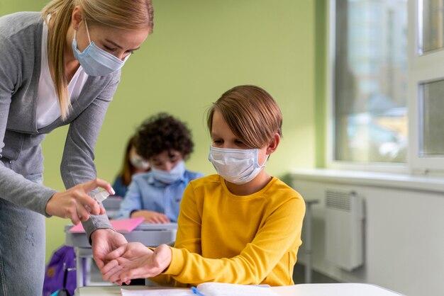 Insegnante femminile con mascherina medica che dà disinfettante per le mani ai bambini in classe