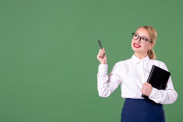 Insegnante femminile di vista frontale che posa con il blocco note e la penna su fondo verde documento dello studente uniforme del libro di studio della scuola del lavoro di lezione della donna