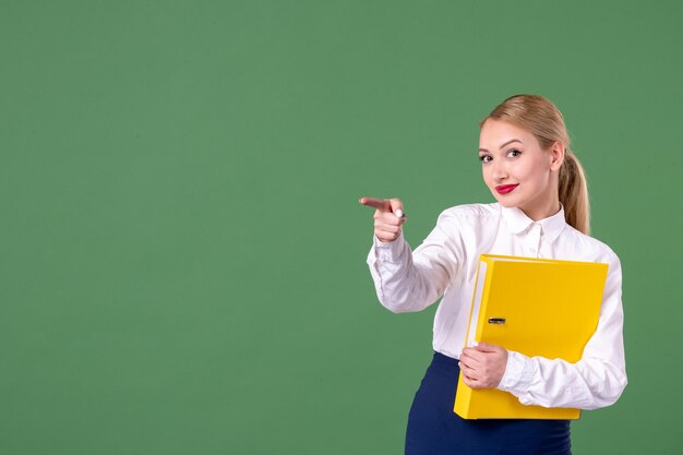 Insegnante femminile di vista frontale che tiene i file gialli e che indica su sfondo verde lezione libro studio biblioteca scuola lavoro uniforme studenti