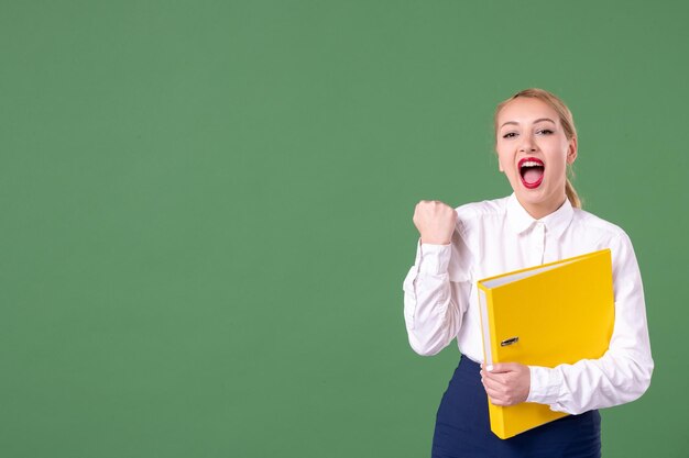 Insegnante femminile di vista frontale in posa con file gialli su sfondo verde libro scuola donna biblioteca studio università lavoro uniforme lezioni per studenti