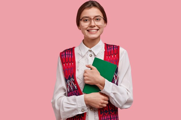 Insegnante femminile soddisfatta in abiti eleganti e ordinati, indossa grandi occhiali ottici, tiene un taccuino verde, felice di incontrare gli alunni dopo le vacanze estive, isolato sul muro rosa. Nerd allegro sulla lezione.