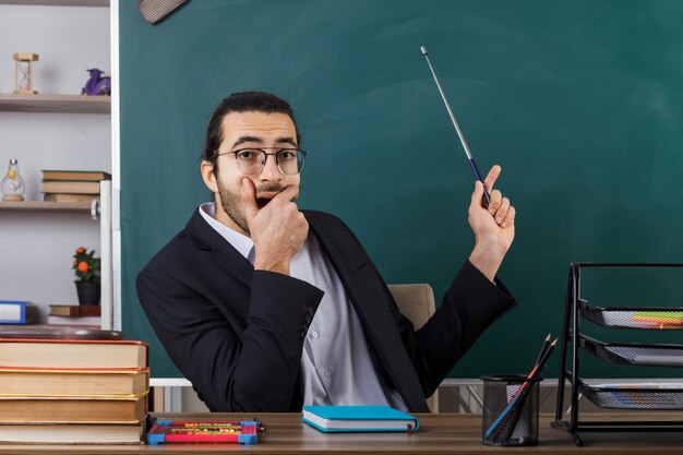 Insegnante maschio con bocca coperta spaventata che indossa occhiali punta con puntatore stick alla lavagna seduto al tavolo con gli strumenti della scuola in classe