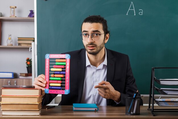 Insegnante maschio con gli occhiali tenendo l'abaco seduto a tavola con gli strumenti della scuola in aula
