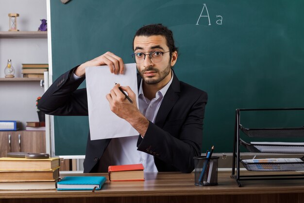Insegnante maschio impressionato con gli occhiali che tiene la carta con la penna seduto al tavolo con gli strumenti della scuola in classe
