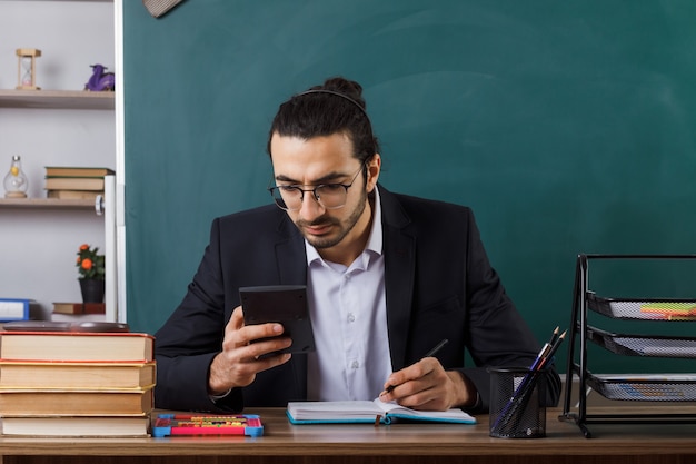 Insegnante maschio pensante con gli occhiali che tiene in mano e guarda la calcolatrice seduto al tavolo con gli strumenti della scuola in classe