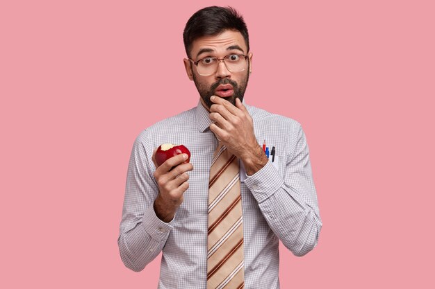 Insegnante stupito con setole spesse tiene il mento, fa uno spuntino tra le lezioni, vestito con una camicia formale con cravatta