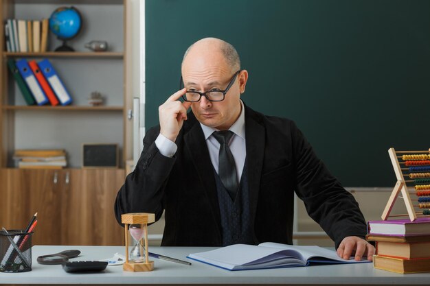 Insegnante uomo che indossa occhiali che controlla il registro di classe con un'espressione seria e sicura seduto al banco della scuola davanti alla lavagna in classe