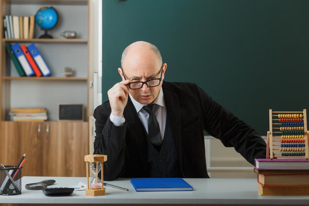 Insegnante uomo che indossa occhiali che controlla il registro di classe guardando la fotocamera con espressione scettica seduto al banco della scuola davanti alla lavagna in classe