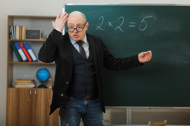 Insegnante uomo con gli occhiali in piedi vicino alla lavagna in classe che spiega la lezione che sembra deluso e confuso