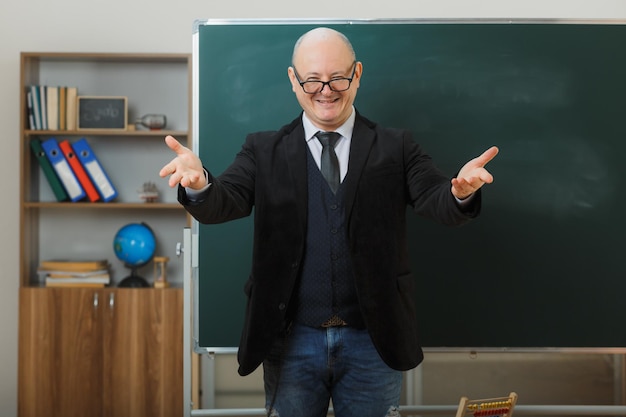 Insegnante uomo con gli occhiali in piedi vicino alla lavagna in classe che spiega la lezione facendo un gesto di benvenuto con le mani sorridenti