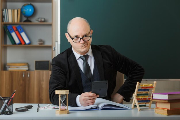 Insegnante uomo con gli occhiali seduto al banco della scuola con il registro di classe davanti alla lavagna in classe spiegando la lezione guardando la calcolatrice scontenta