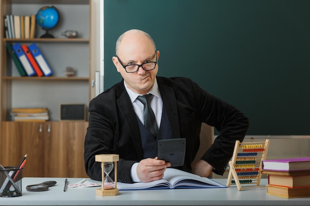 Insegnante uomo con gli occhiali seduto al banco della scuola con il registro di classe davanti alla lavagna in classe spiegando la lezione guardando la calcolatrice scontenta