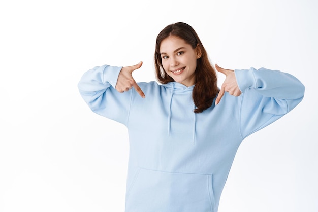 Inserisci qui il testo del tuo logo Giovane donna sorridente in felpa con cappuccio che punta il dito al centro o verso il basso mostrando pubblicità che dimostra il tuo banner promozionale in piedi su sfondo bianco