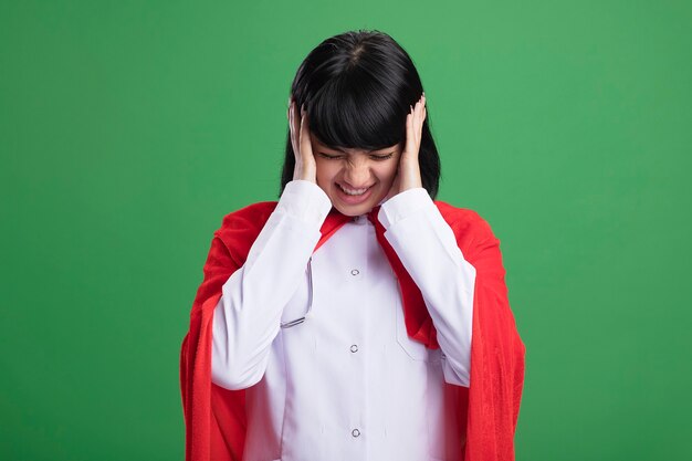 Insoddisfatto con la testa abbassata giovane ragazza del supereroe che indossa uno stetoscopio con veste medica e mantello che mette le mani sulle orecchie isolate su verde