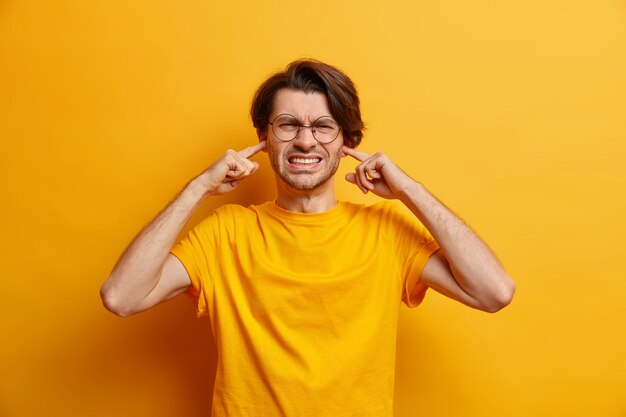 insoddisfatto uomo europeo stringe i denti non riesce a concentrarsi a causa del rumore indossa occhiali maglietta casual sente urlo straziante isolato sul muro giallo. Non volendo ascoltare